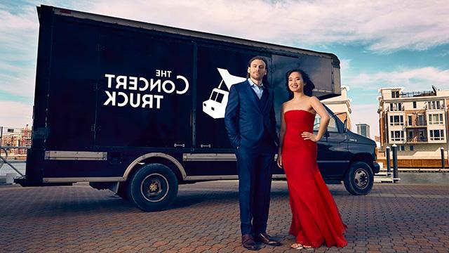 一个穿红裙子的女人和一个穿西装的男人站在一辆黑色卡车前，卡车上写着“音乐会卡车”